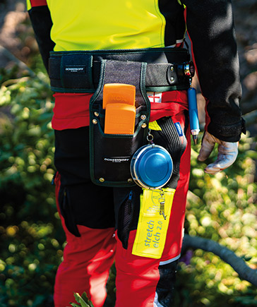 Nahaufnahme eines Forstarbeiters mit Werkzeuggrtel, an dem Keile, Maband, Spray und weitere Arbeitsmittel befestigt sind.