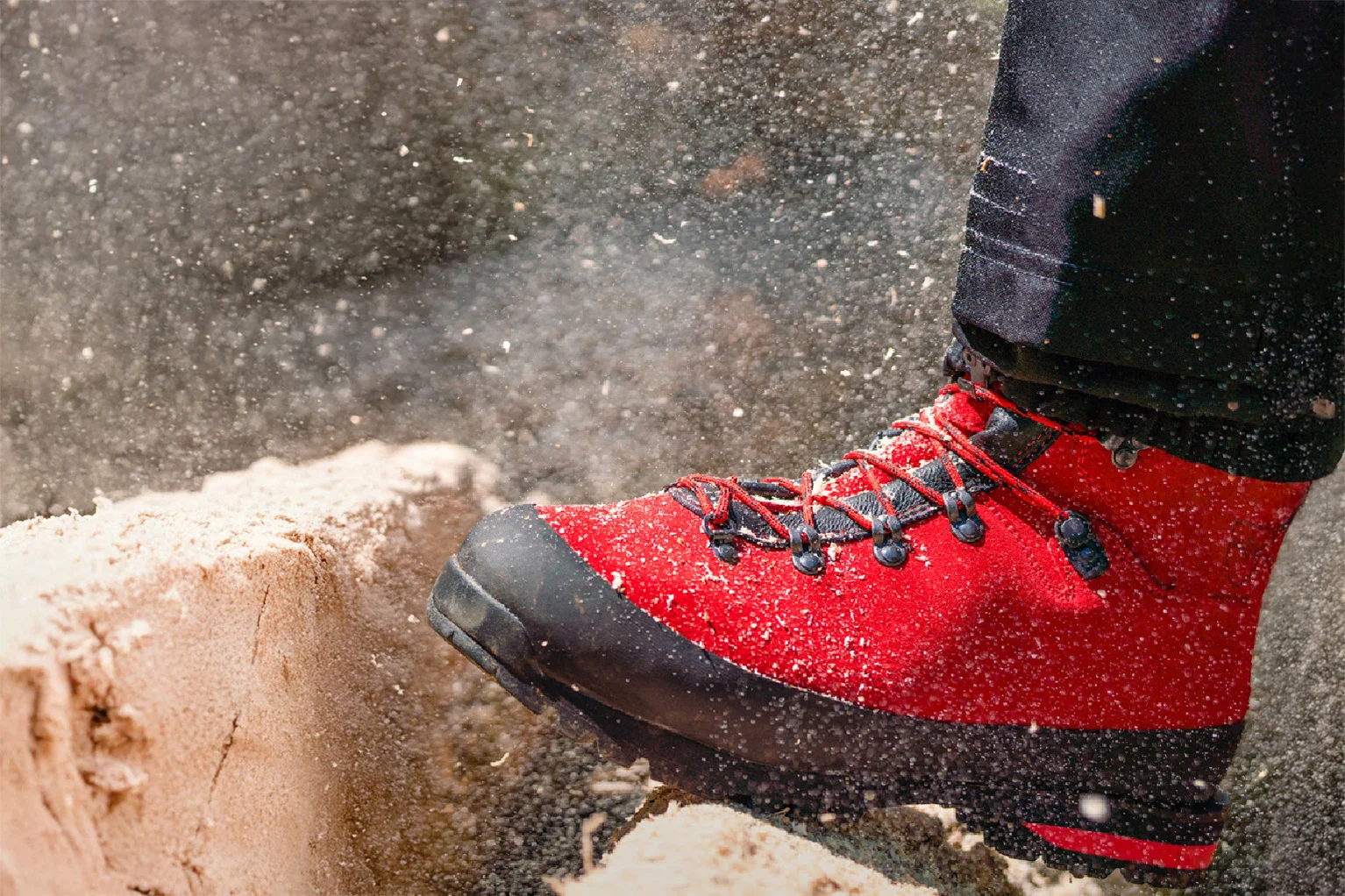 Nahaufnahme eines roten Forststiefels mit schwarzer Schutzkappe und Schn�rung. Der Schuh steht auf einem frisch ges�gten Baumstumpf, Holzsp�ne fliegen durch die Luft und betonen die Arbeitssituation im Wald.