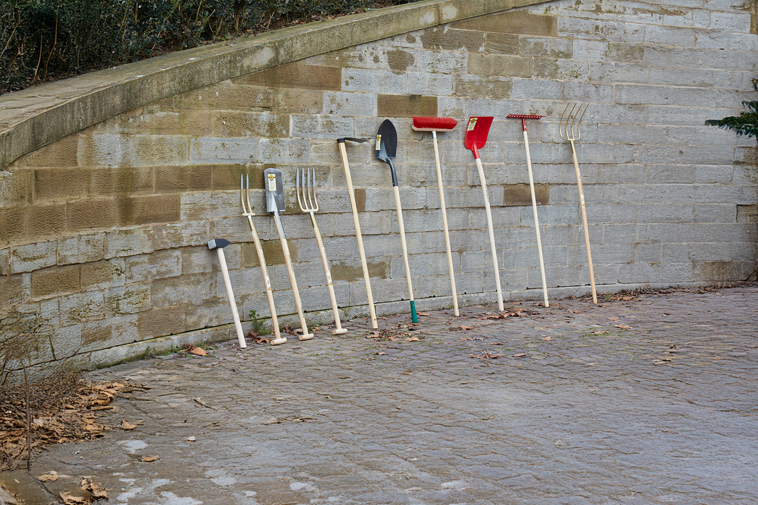 Mehrere Gartenger�te mit Holzstielen lehnen an einer Steinmauer, darunter Hammer, Grabegabeln, Spaten, Besen, Schneeschieber, Rechen und Mistgabel.